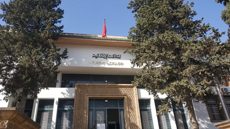 Un bâtiment blanc avec une grande porte dorée. L'image est encadrée par deux arbres. On voit un intitulé en arabe et un deuxième en Tamazight.