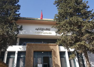Un bâtiment blanc avec une grande porte dorée. L'image est encadrée par deux arbres. On voit un intitulé en arabe et un deuxième en Tamazight.